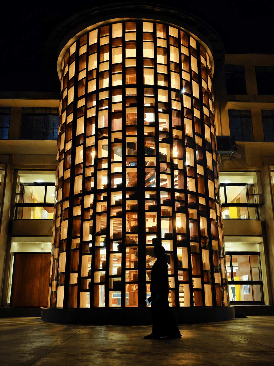 The Devagiri Library at night: a tall cylindrical reading tower in a black steel grid, lit from within, with a figure walking past at ground level